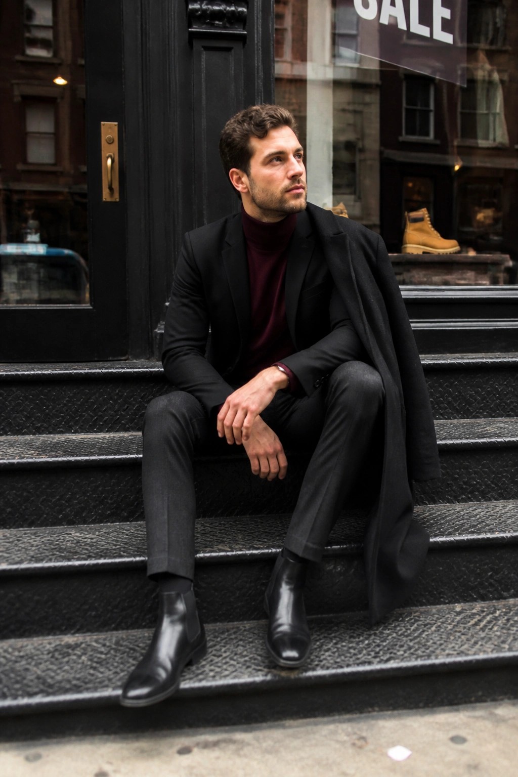 Man in black suit over maroon turtleneck seated on stone steps