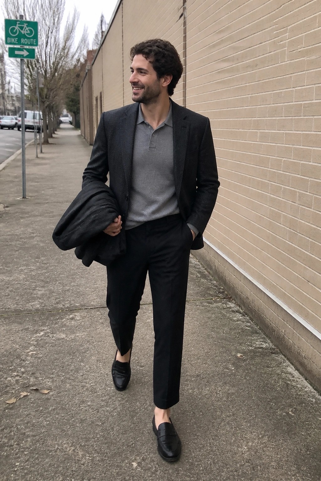 Man in dark patterned blazer over gray polo shirt, black trousers, and black loafers while holding a jacket on a sidewalk.