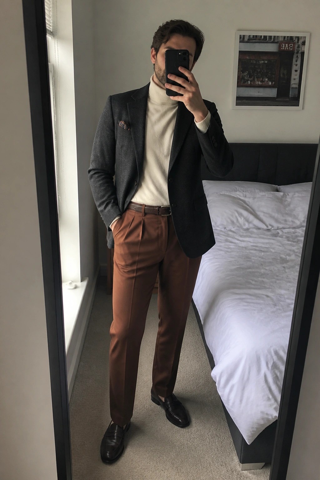 Man in gray blazer and cream turtleneck taking mirror selfie in bedroom