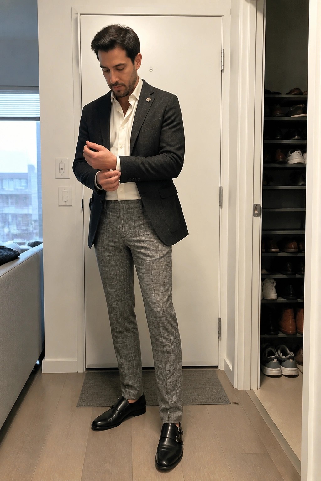Man wearing dark gray blazer, white dress shirt, gray plaid trousers, and black double monk strap shoes while adjusting cuff.