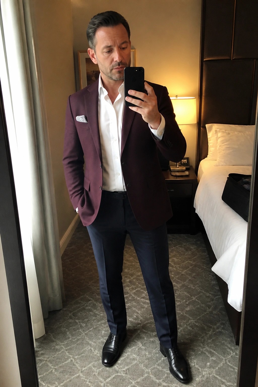 Man in maroon blazer and navy pants taking mirror selfie in hotel room