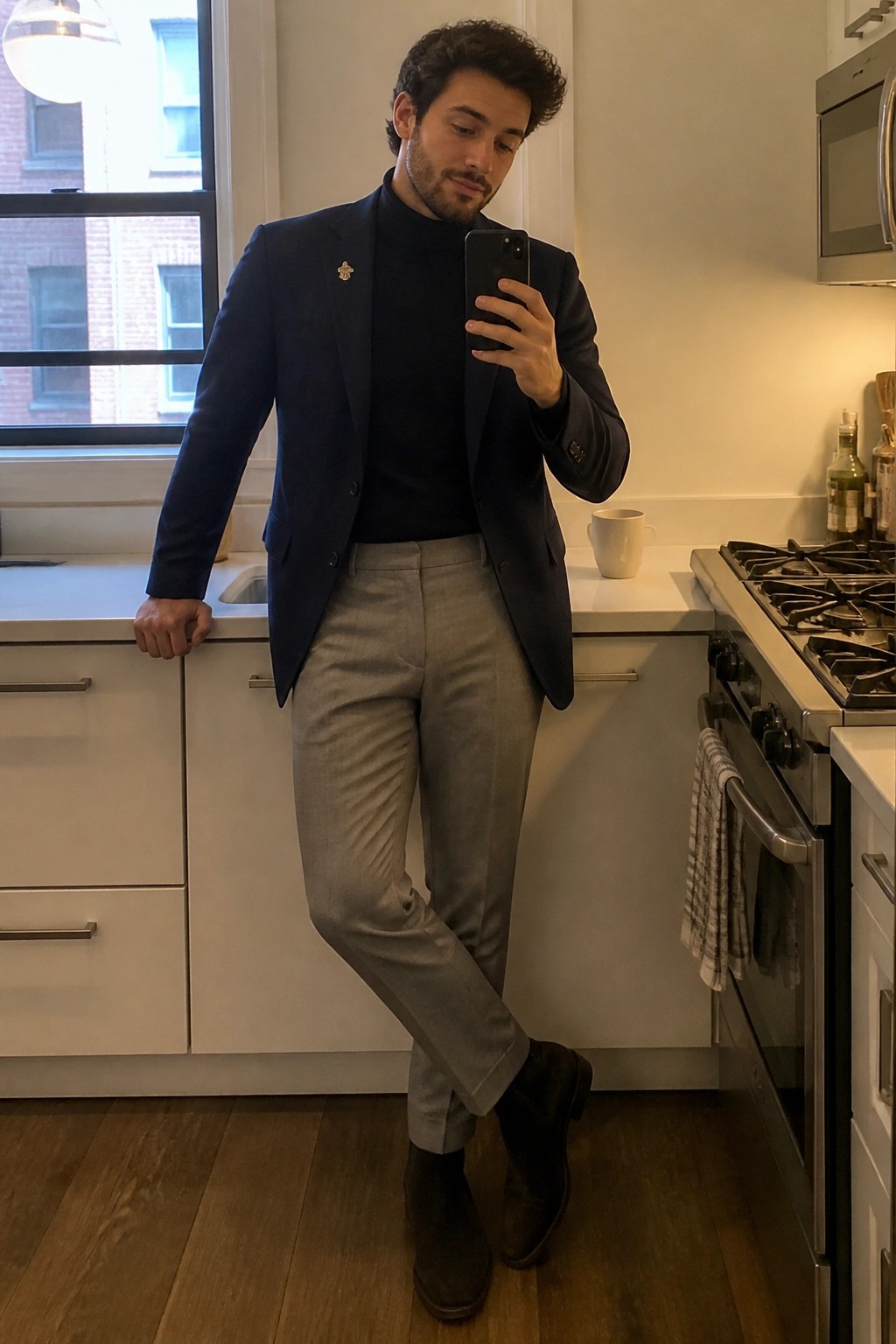 Man in navy blazer over black turtleneck with gray pants taking kitchen selfie.