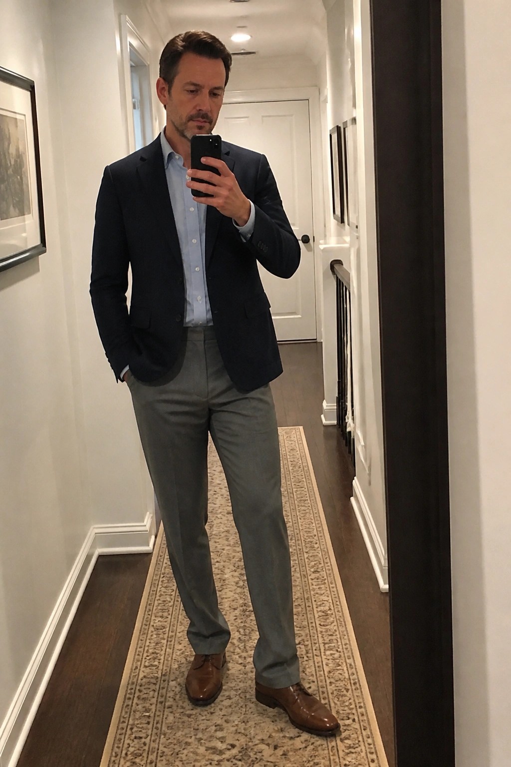 Man wearing a navy blazer over a light blue button down shirt with gray dress trousers and brown leather shoes while taking a mirror selfie in a hallway.