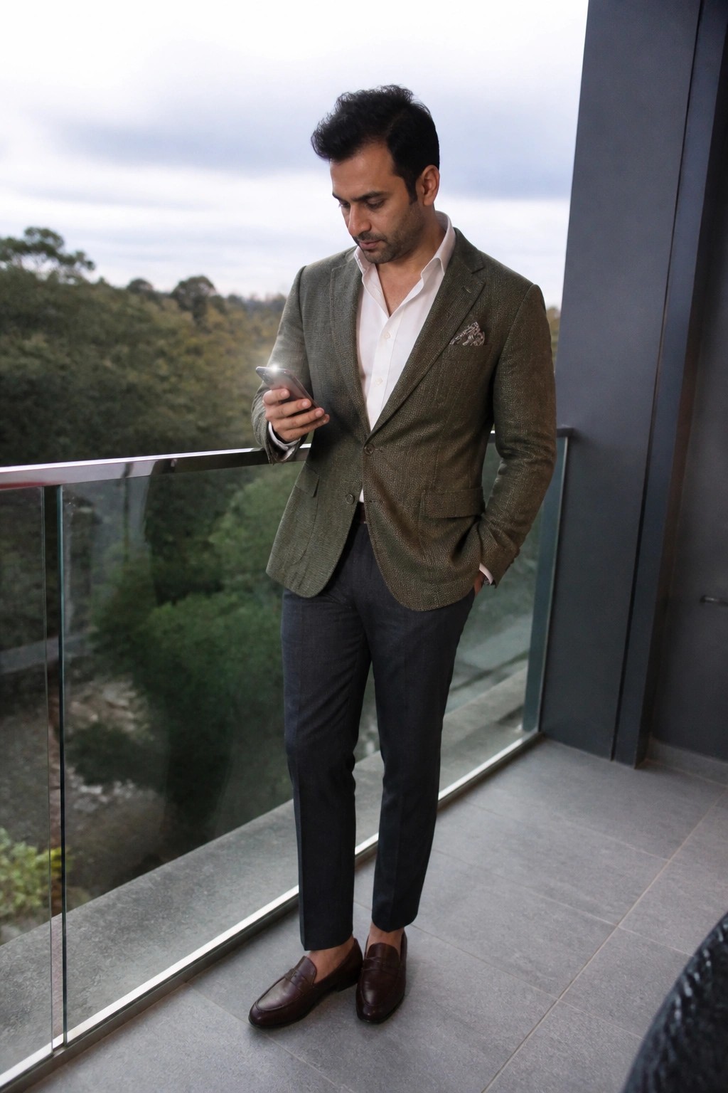 Man in green blazer and white shirt checks smartphone on balcony with trees below