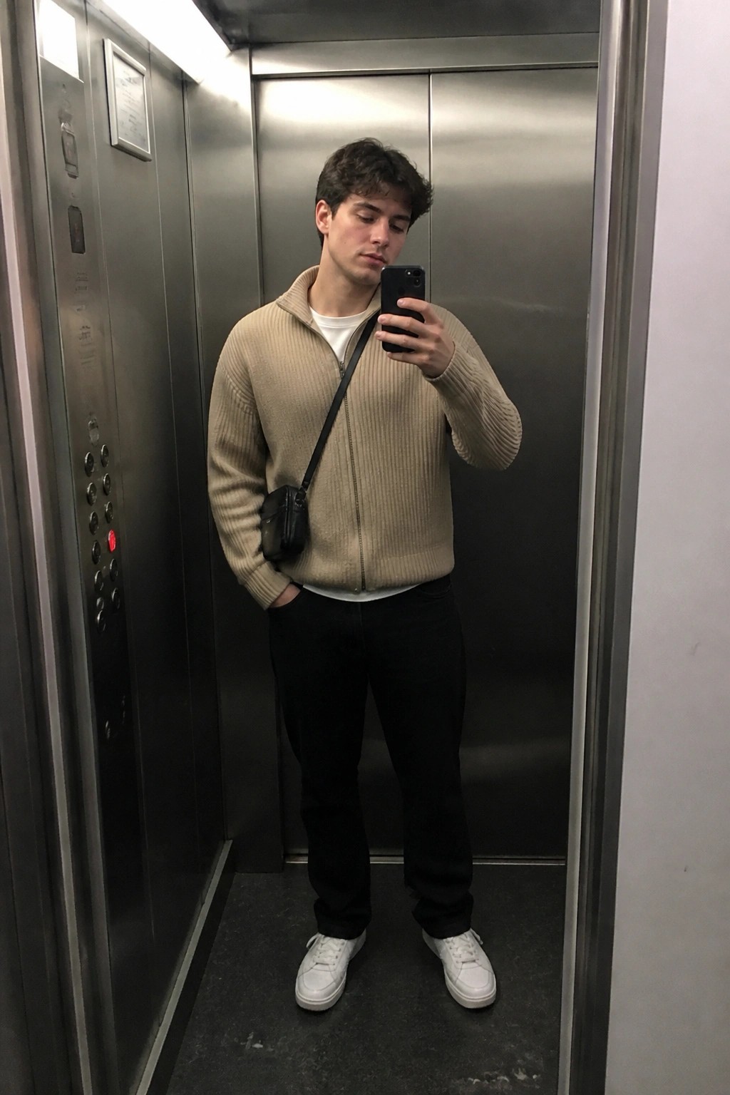 Man in beige zip-up sweater with black bag taking selfie in elevator