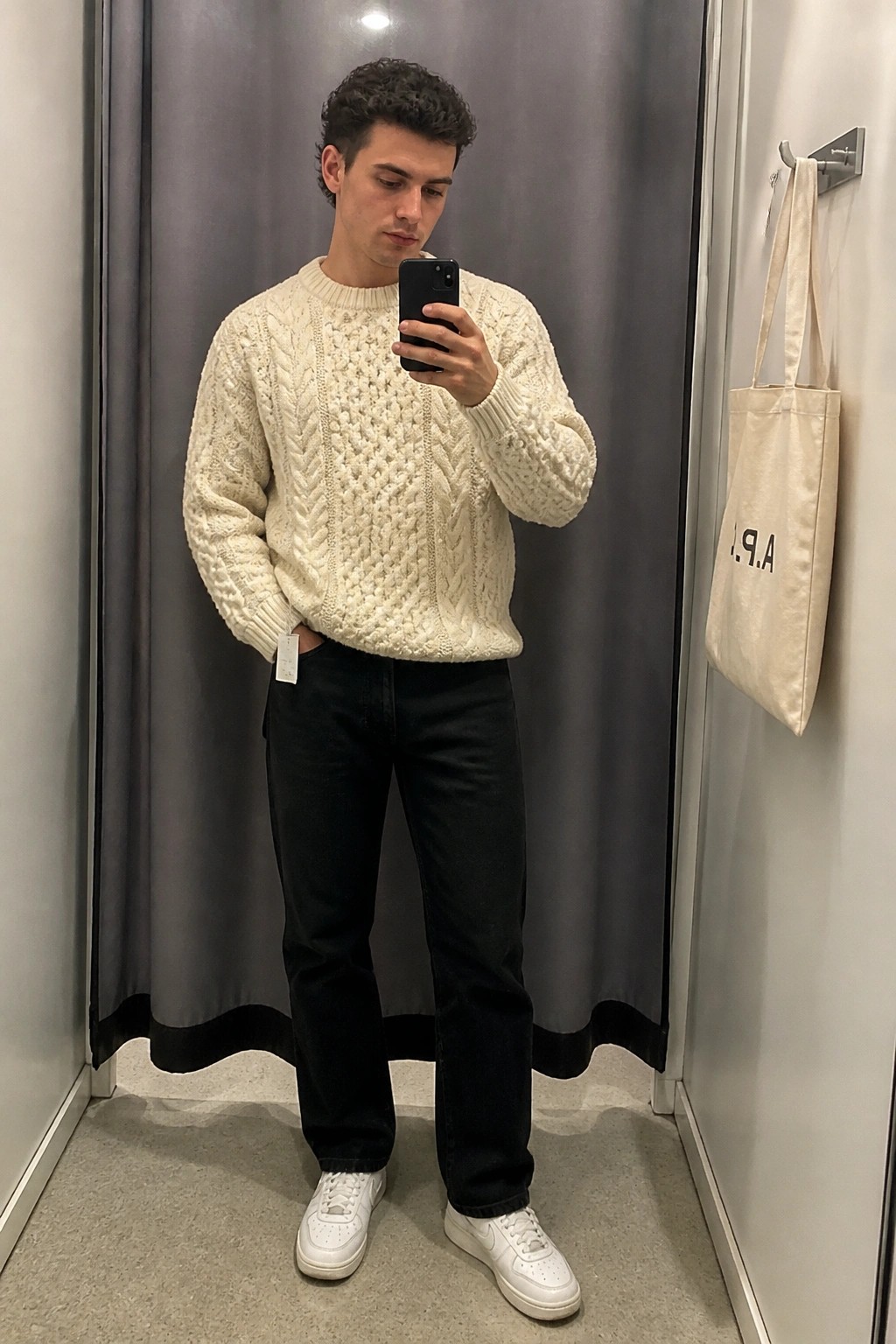 Man in cream cable-knit sweater and black jeans taking mirror selfie in fitting room.