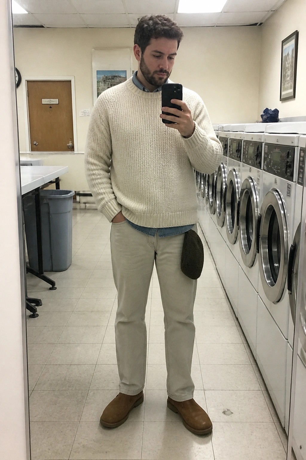 Man in cream knit sweater and khaki pants taking mirror selfie in laundromat.