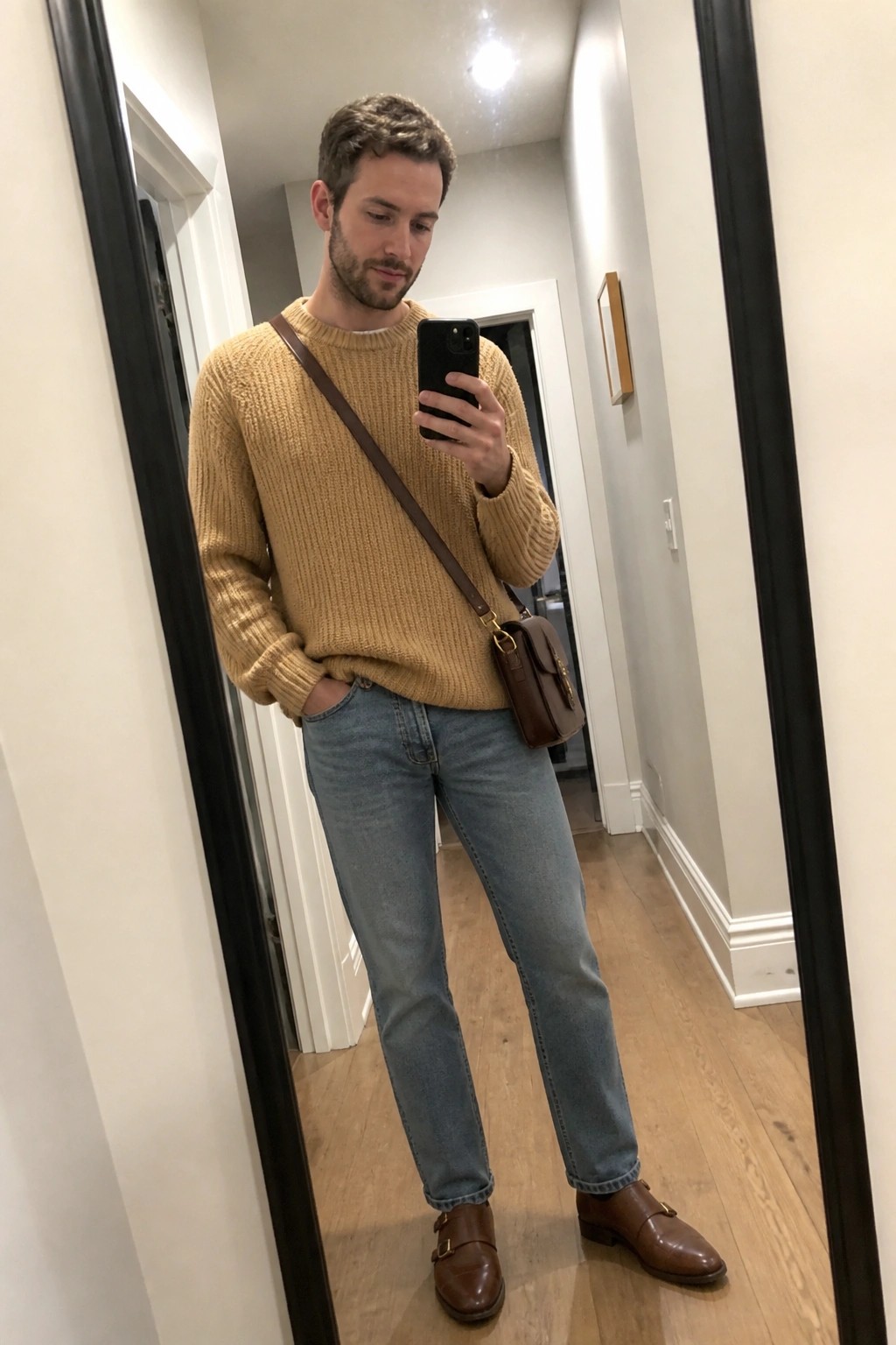 Man in tan knit sweater and blue jeans with brown leather crossbody bag in hallway
