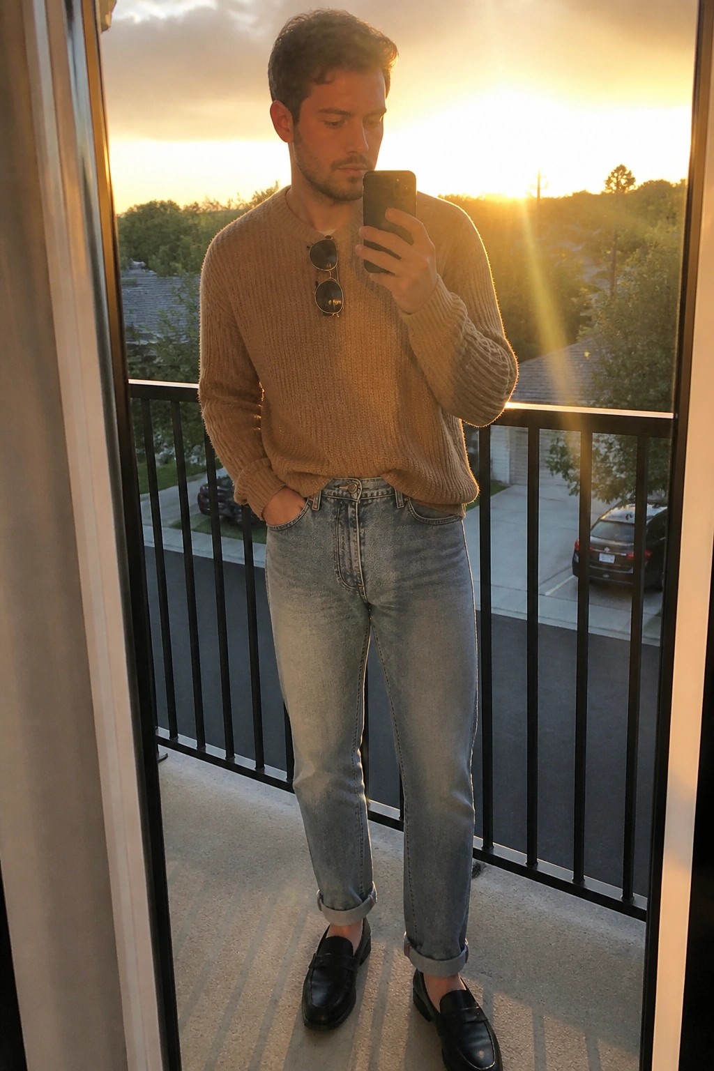 Man in tan sweater and cuffed jeans taking phone selfie on sunset balcony