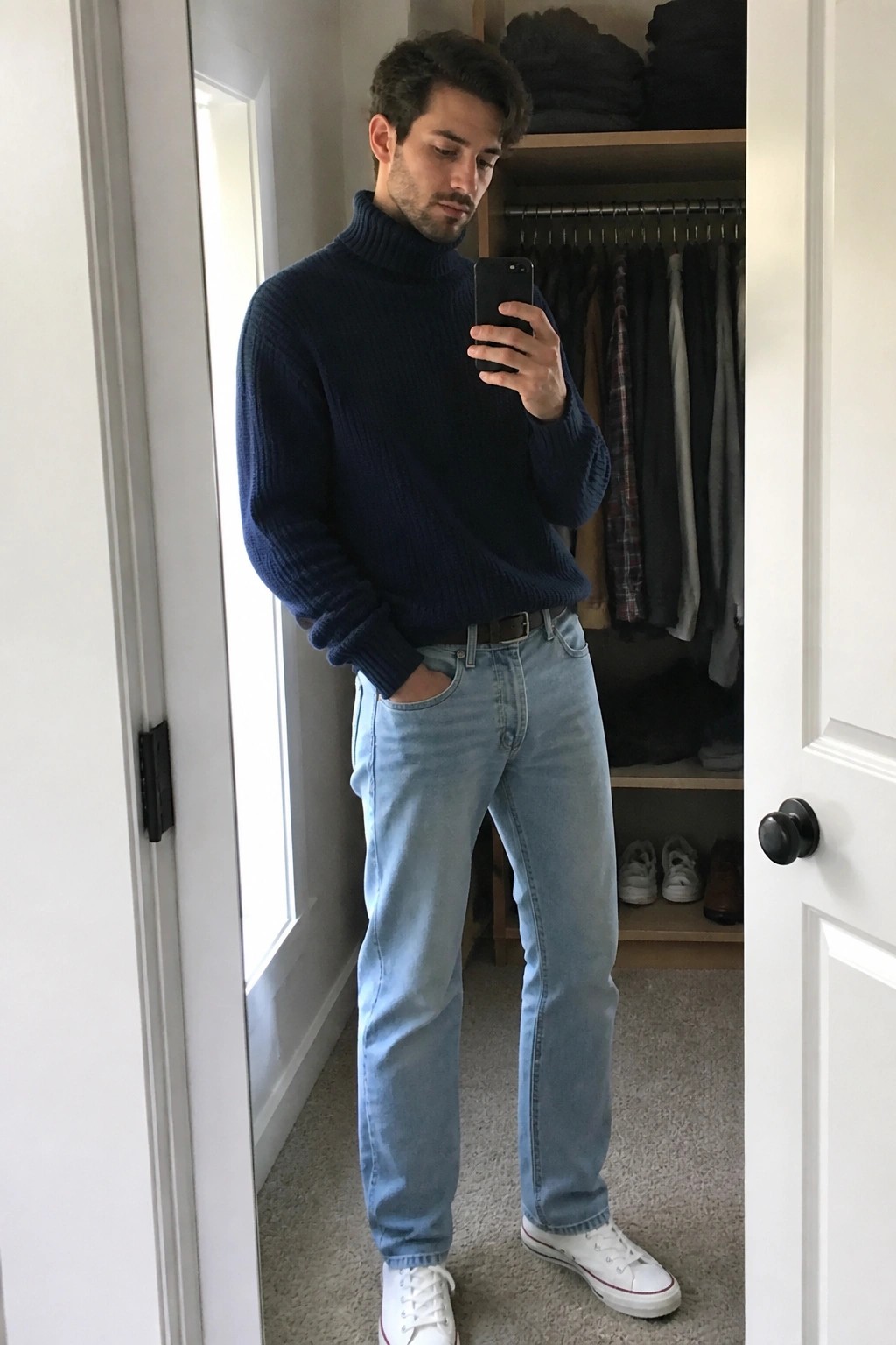 A man wearing a navy chunky ribbed turtleneck sweater tucked slightly into light blue straight leg jeans with a brown belt, white canvas sneakers, standing in a closet taking a mirror selfie.