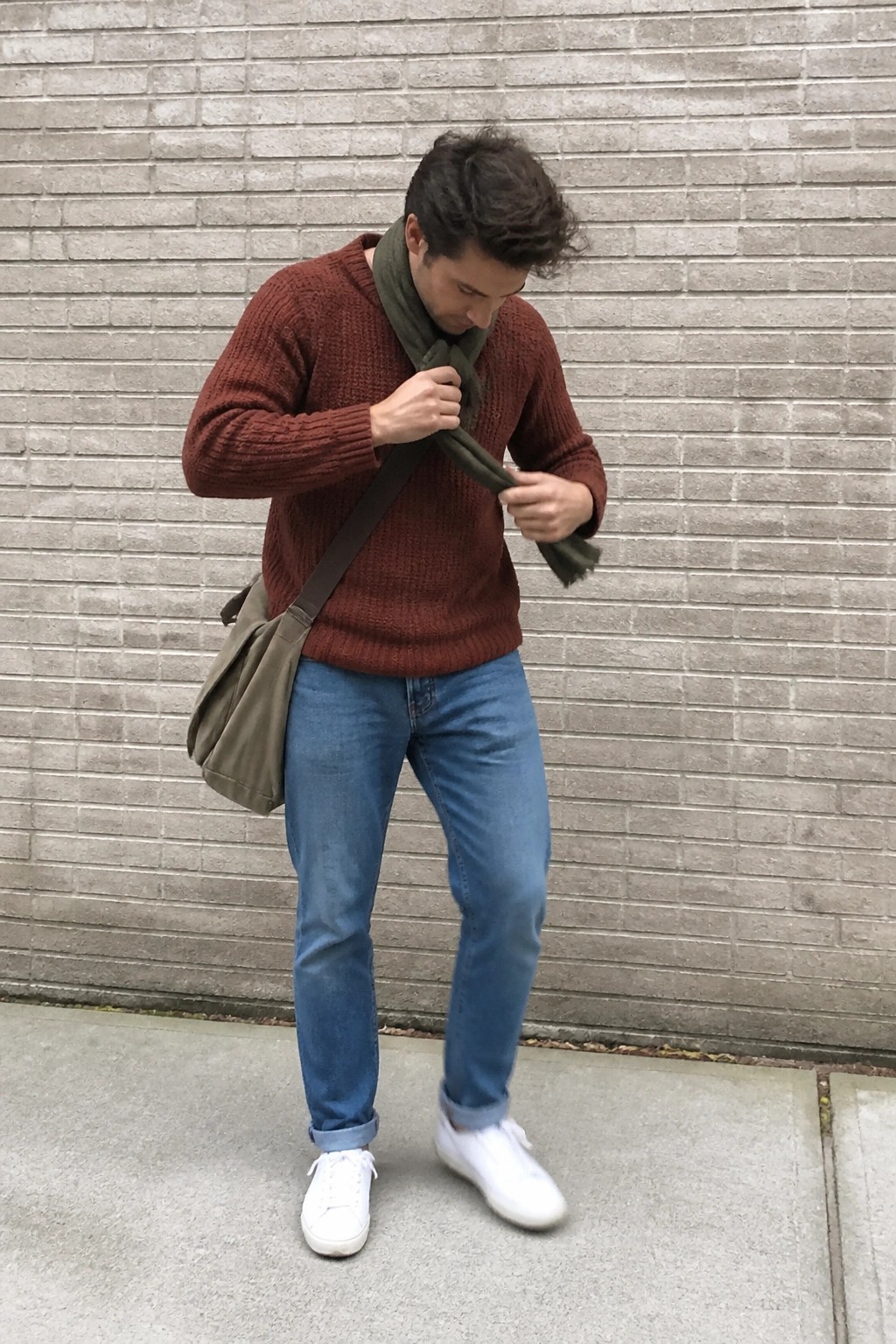 Man in rust sweater adjusts green scarf with messenger bag by brick wall