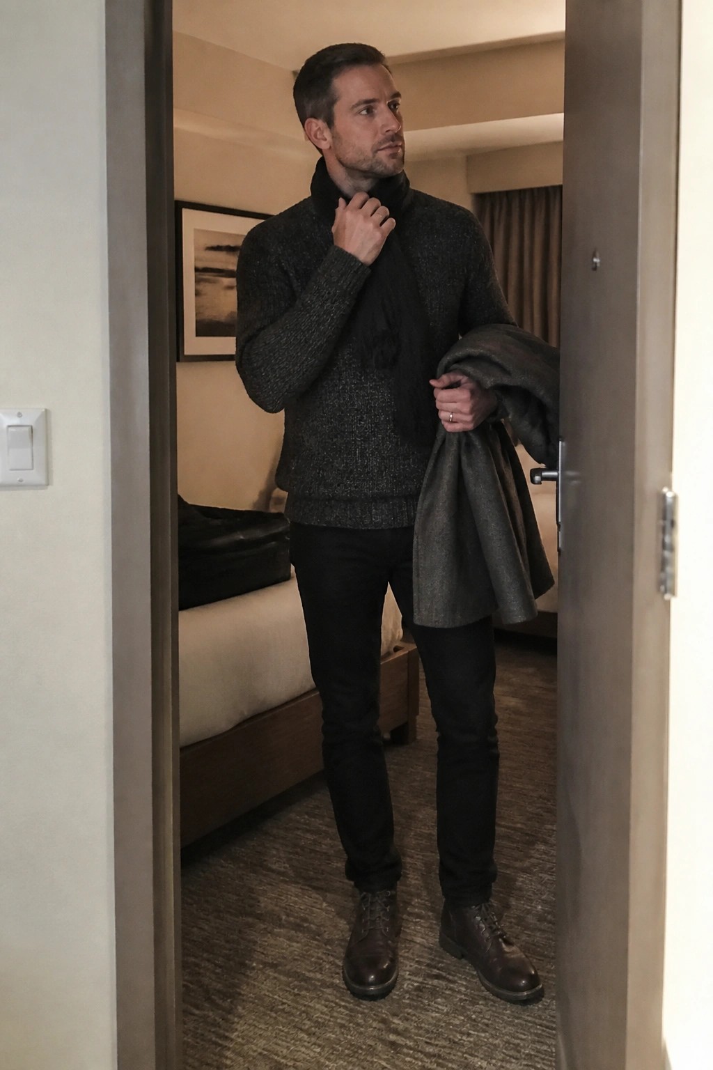Man in dark chunky knit sweater with black scarf black straight leg jeans and brown leather boots holding a gray coat in doorway