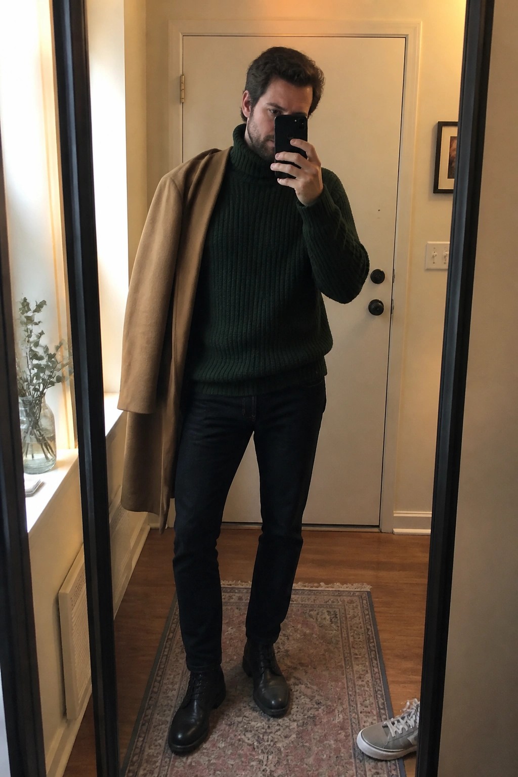 Man in green turtleneck with tan coat over shoulder taking mirror selfie in hallway