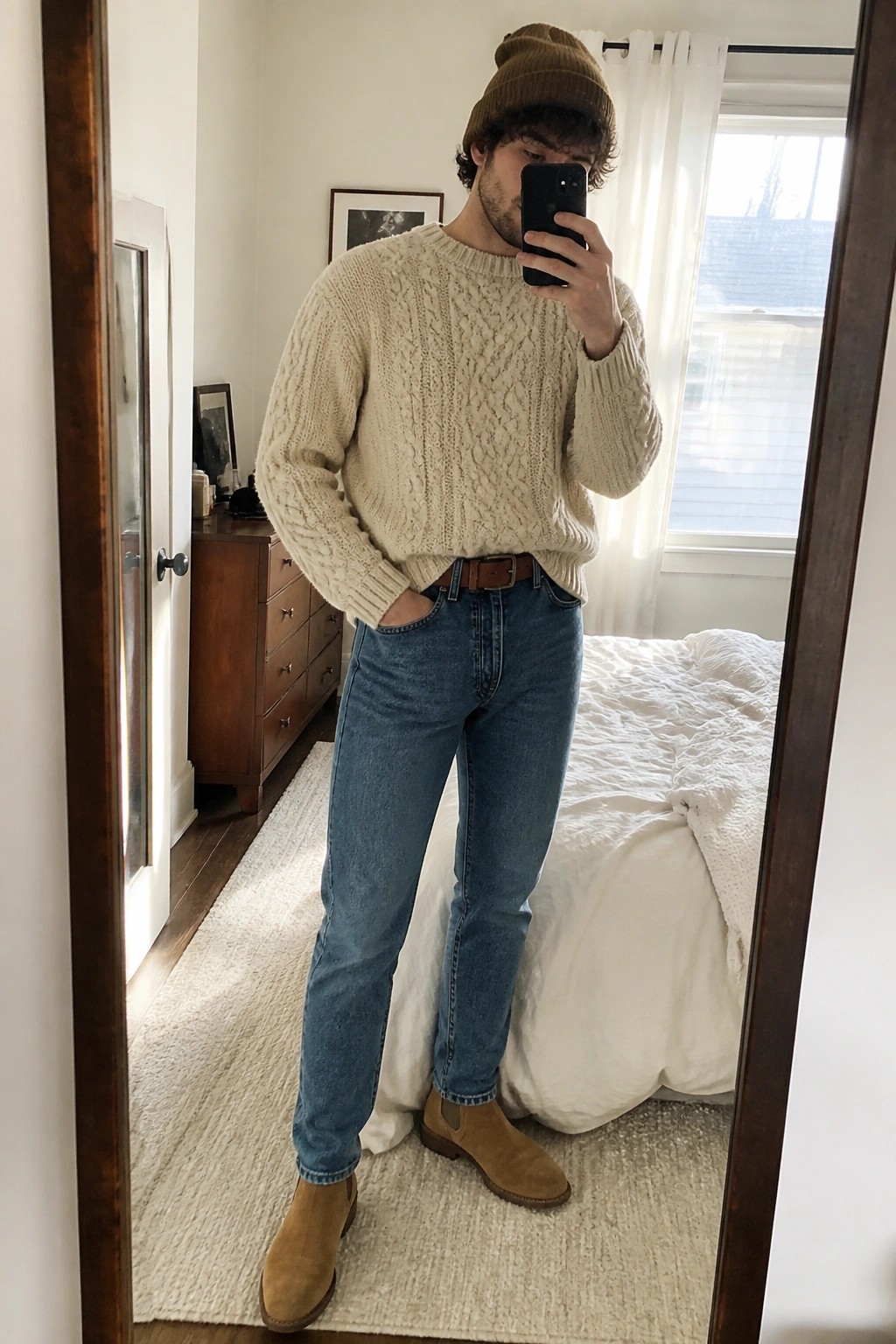 Man in cream cable-knit sweater, blue jeans, brown beanie and boots taking mirror selfie.