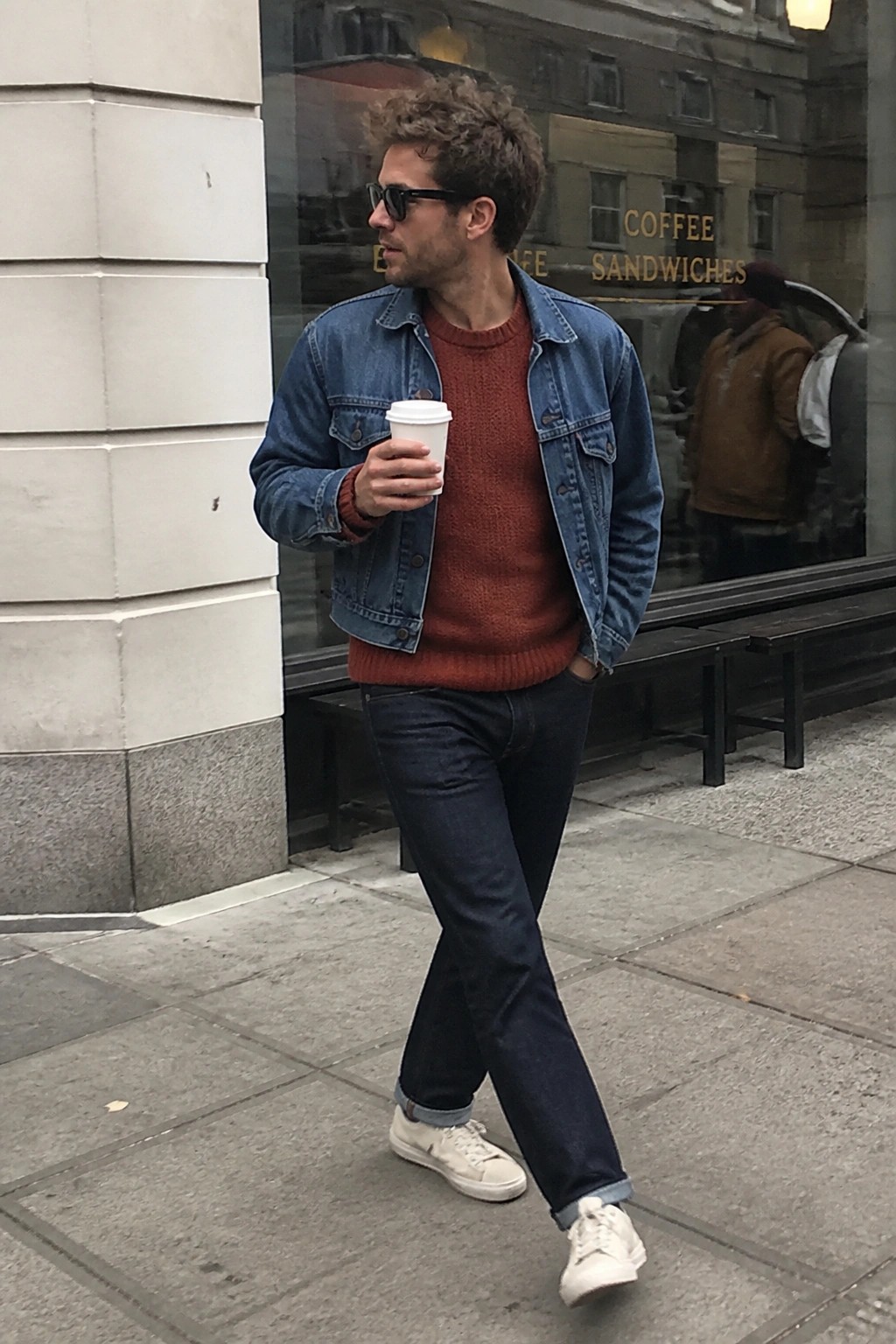 Man in denim jacket over rust sweater holds coffee while walking on sidewalk.