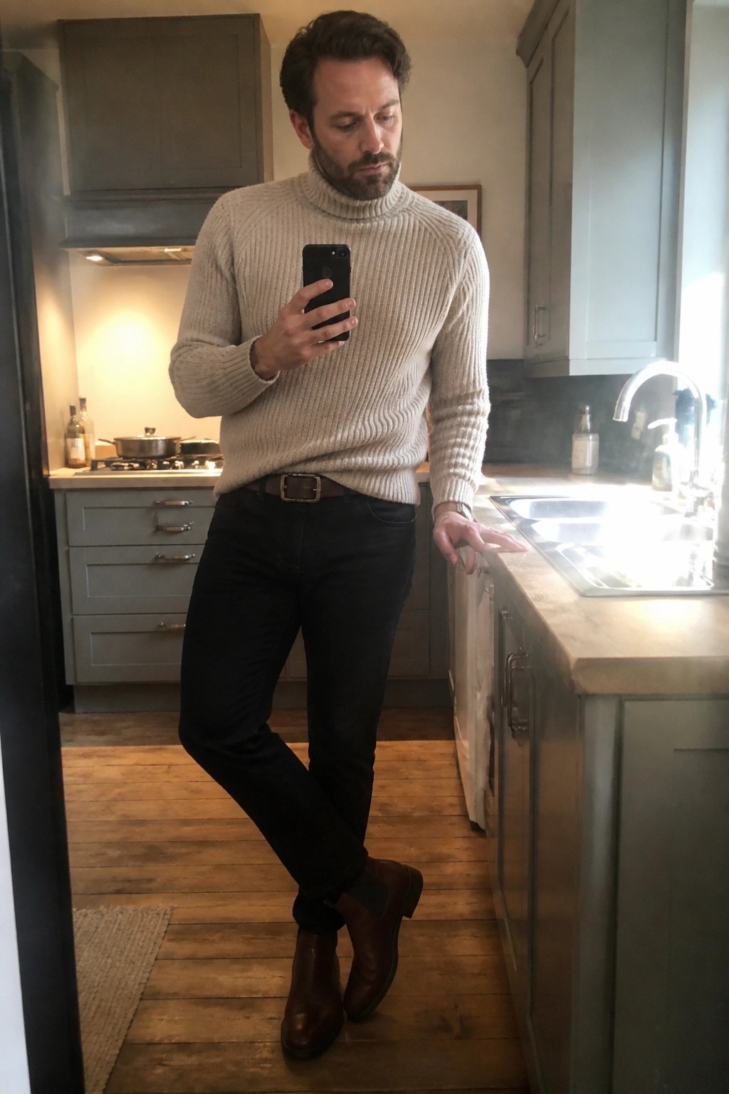 Man in beige turtleneck sweater, dark jeans, and brown boots taking kitchen selfie