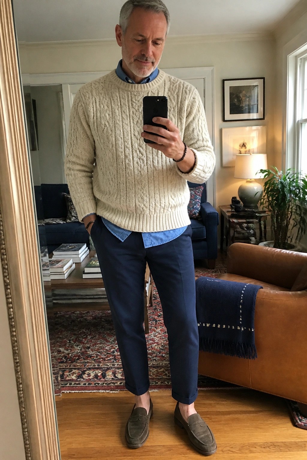 Gray-haired man in cream cable-knit sweater and navy pants taking mirror selfie