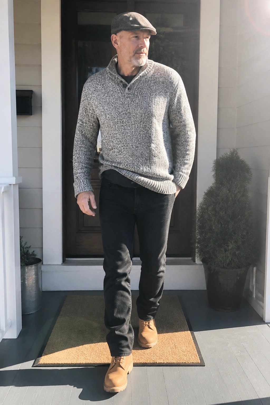 Man in gray marled sweater, flat cap, black jeans, and boots on porch.