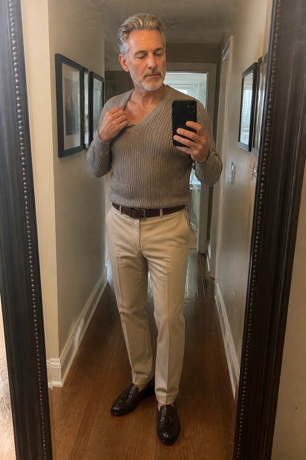 Man in gray V-neck sweater and beige pants taking mirror selfie in hallway