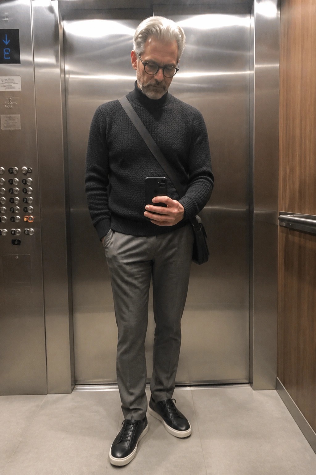 Gray-haired man in textured turtleneck sweater checks phone in elevator