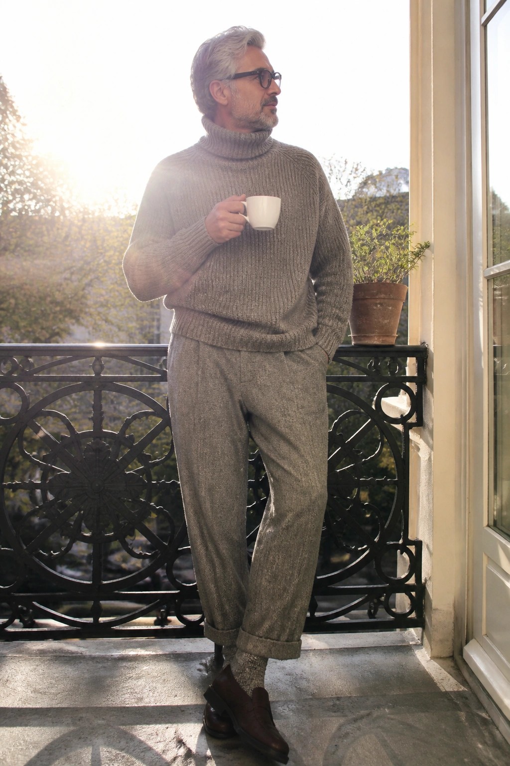 Man in gray turtleneck sweater and pants holding mug on sunny balcony