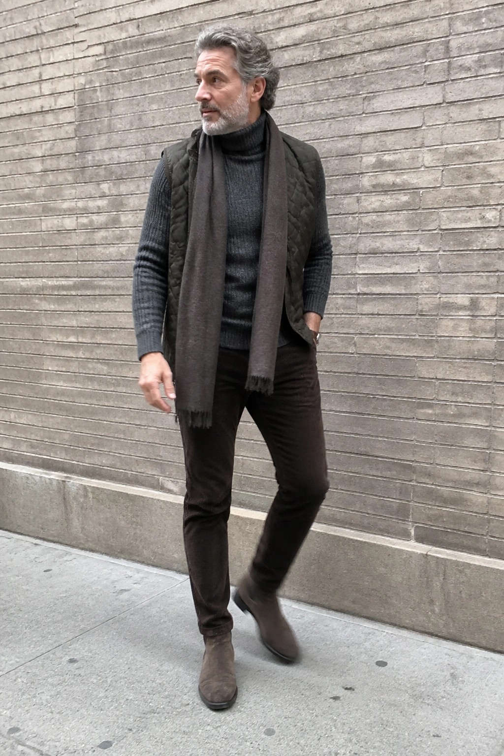 Man over 50 in gray textured turtleneck sweater layered under dark green quilted vest with long brown scarf brown pants and brown suede boots standing against brick wall.