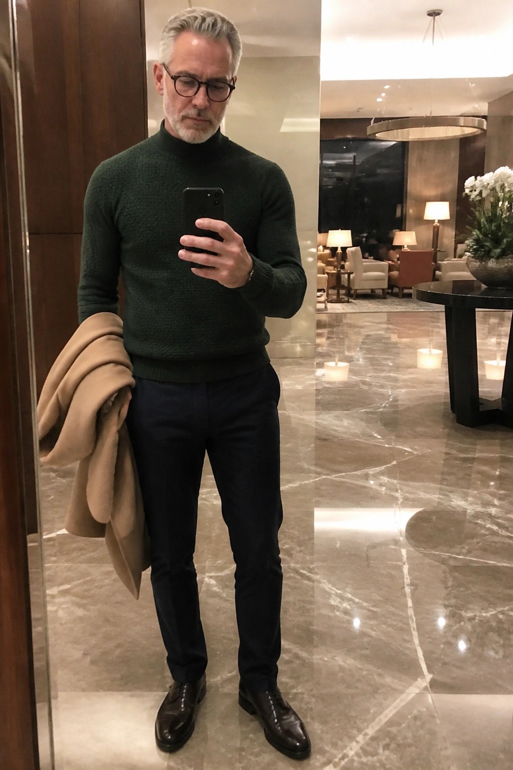 Man in green turtleneck sweater holds tan coat and phone in marble lobby