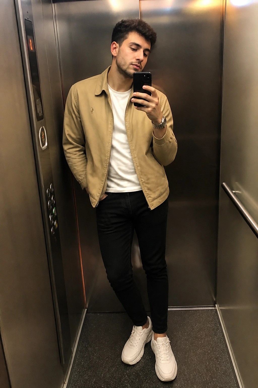 Man wearing beige zip jacket over white t-shirt with black jeans and white sneakers taking a mirror selfie inside an elevator.