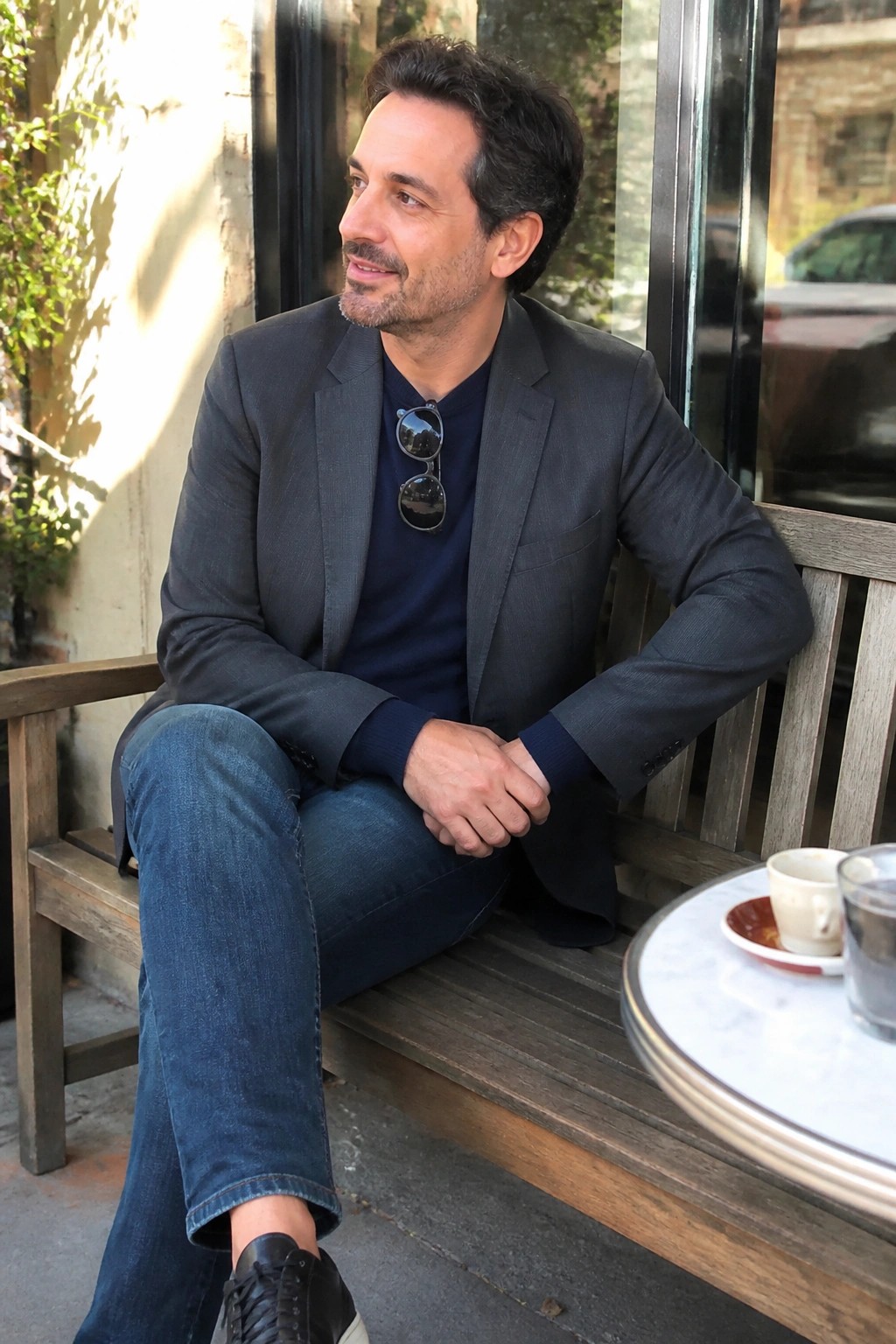 Man in gray blazer and blue jeans sits on wooden cafe bench with coffee