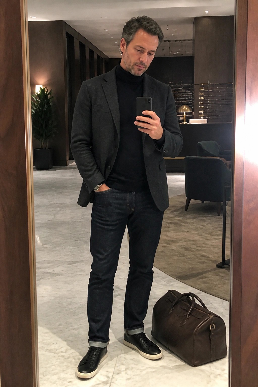 Man in dark blazer and turtleneck taking mirror selfie beside brown leather bag