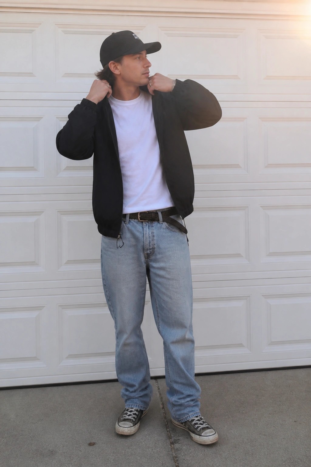 Young man in black bomber jacket adjusts collar before white garage door.