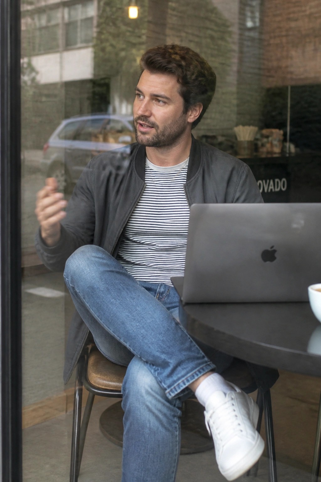 Man in gray bomber jacket and striped shirt gesturing at cafe laptop table