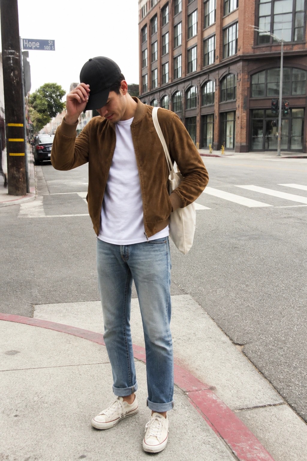 Man in brown suede jacket adjusts black cap on city street with tote bag