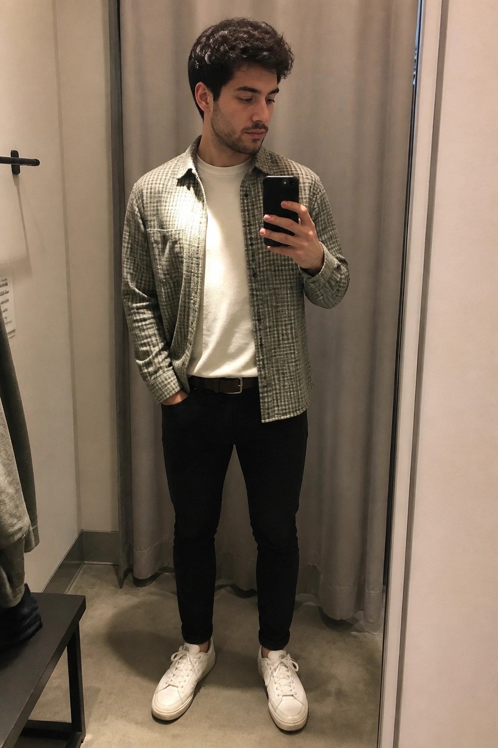 Man in checkered shirt and black pants takes mirror selfie in fitting room