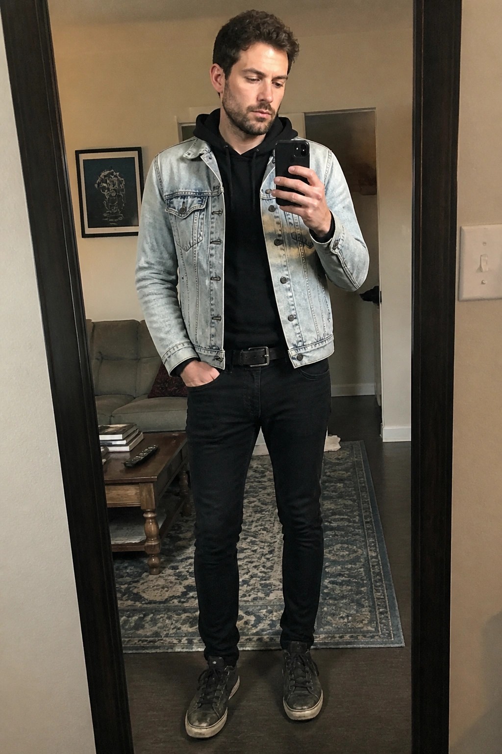 Man in light denim jacket over black hoodie holding phone in mirror reflection
