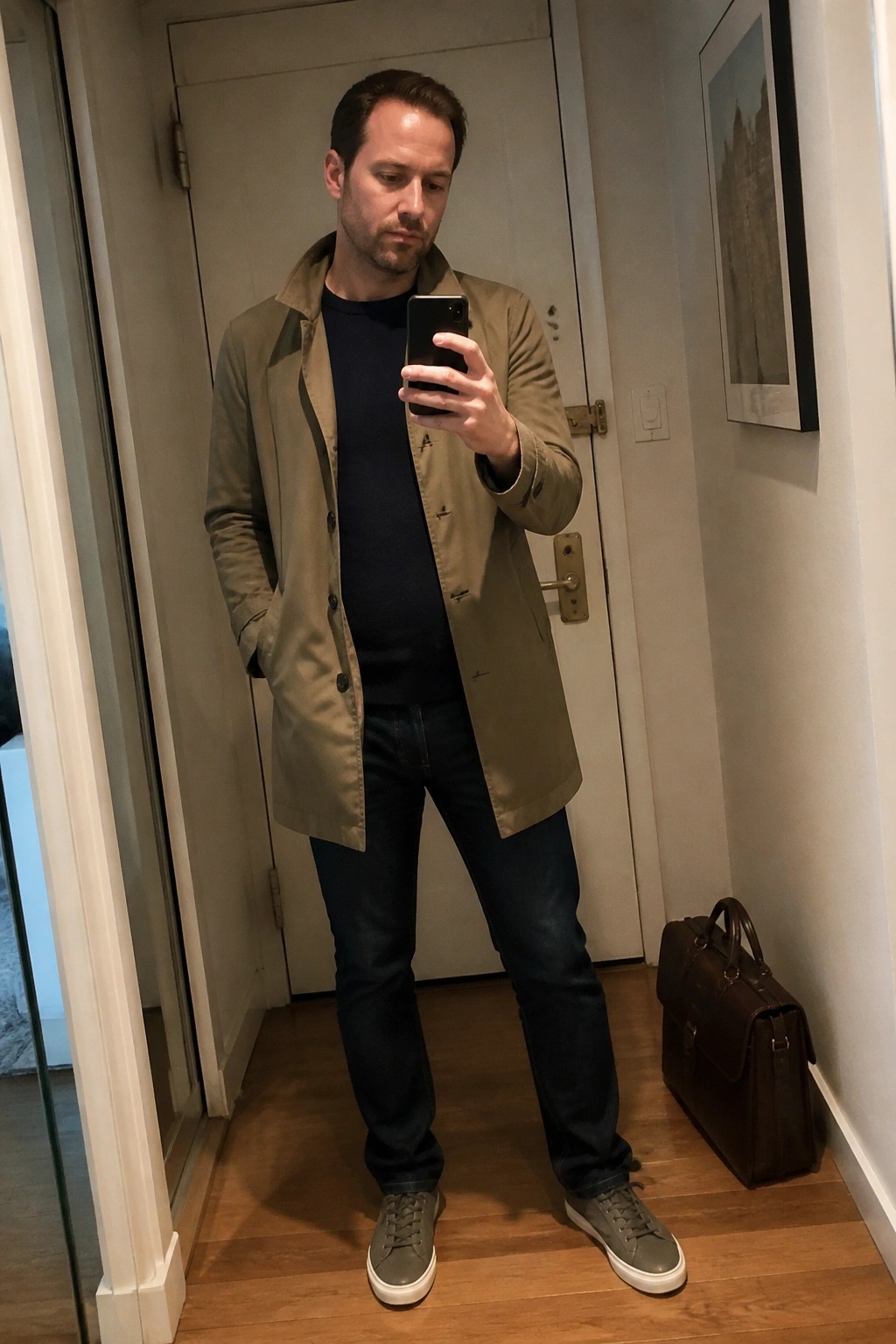 Man in olive coat and jeans taking mirror selfie in hallway with leather bag nearby