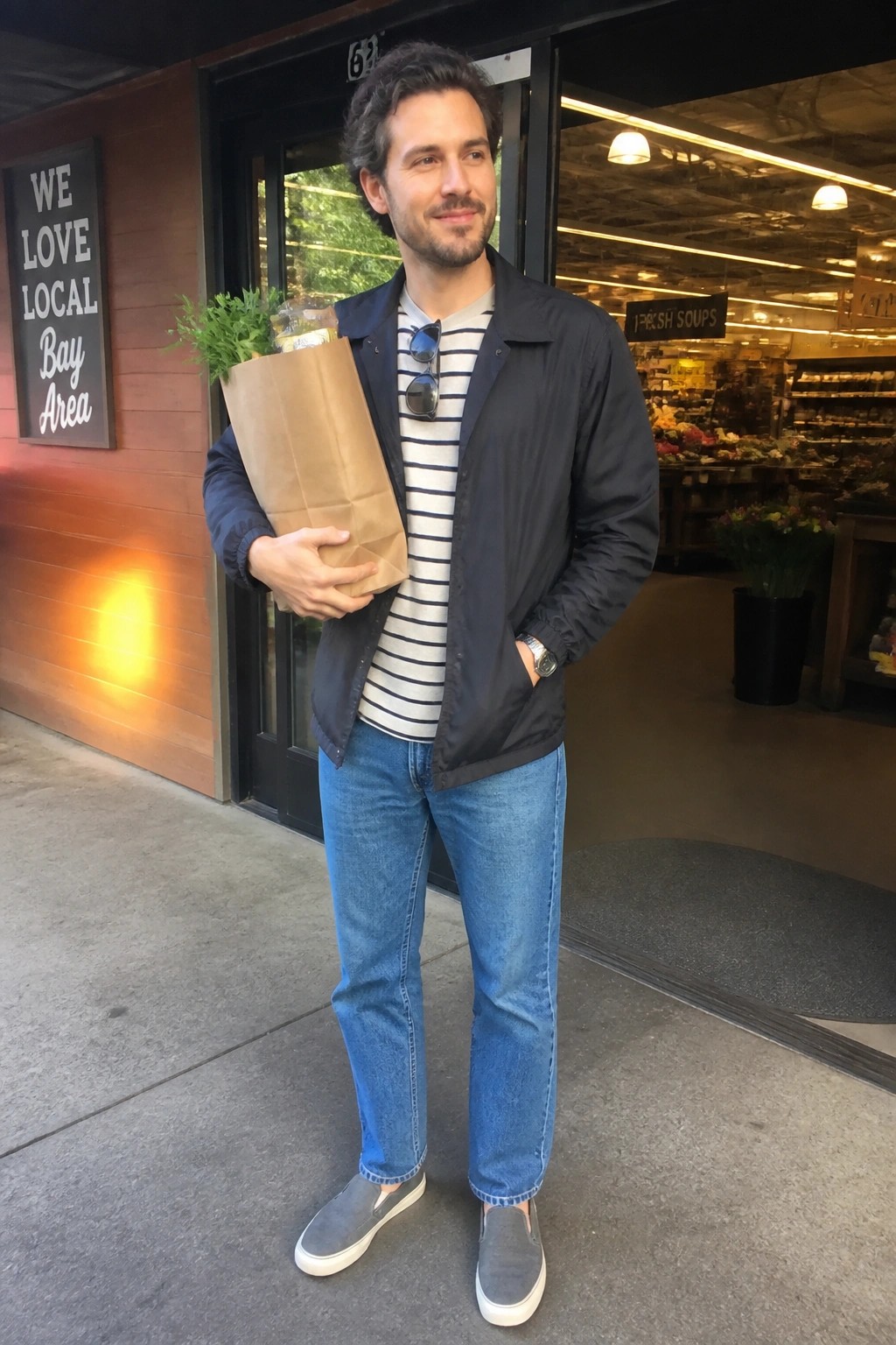 Man in navy jacket over striped shirt holds paper bag outside market