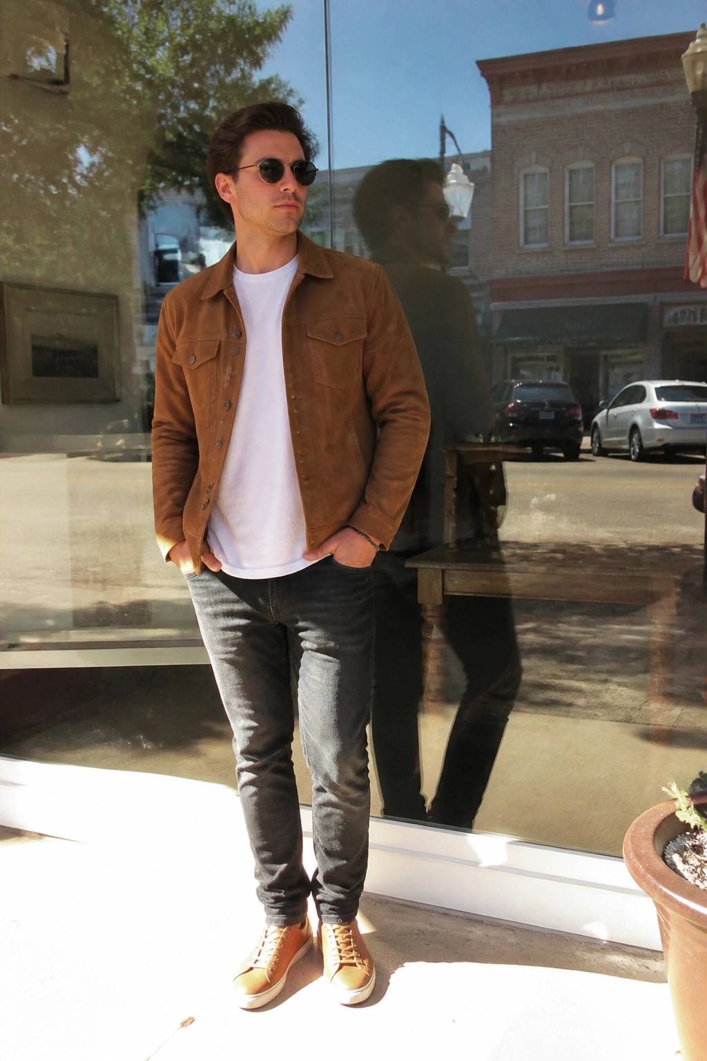 Man in brown suede jacket and jeans stands before reflective storefront window on sunny street