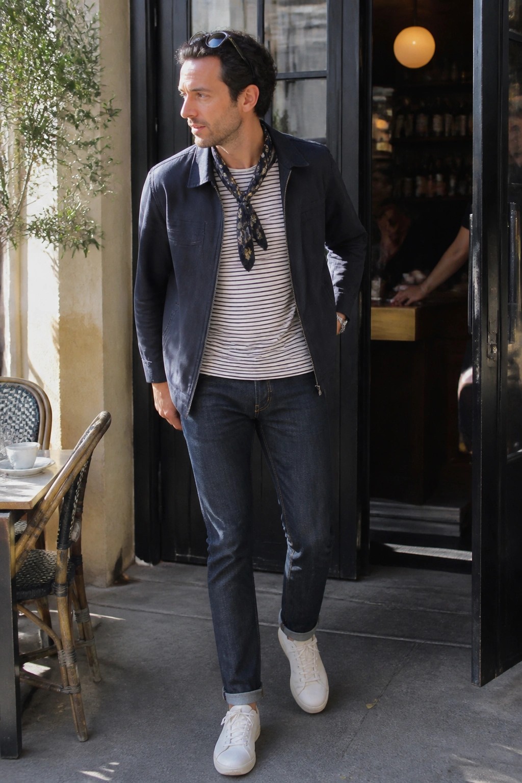 Man in navy jacket, striped shirt, scarf, and jeans at cafe entrance