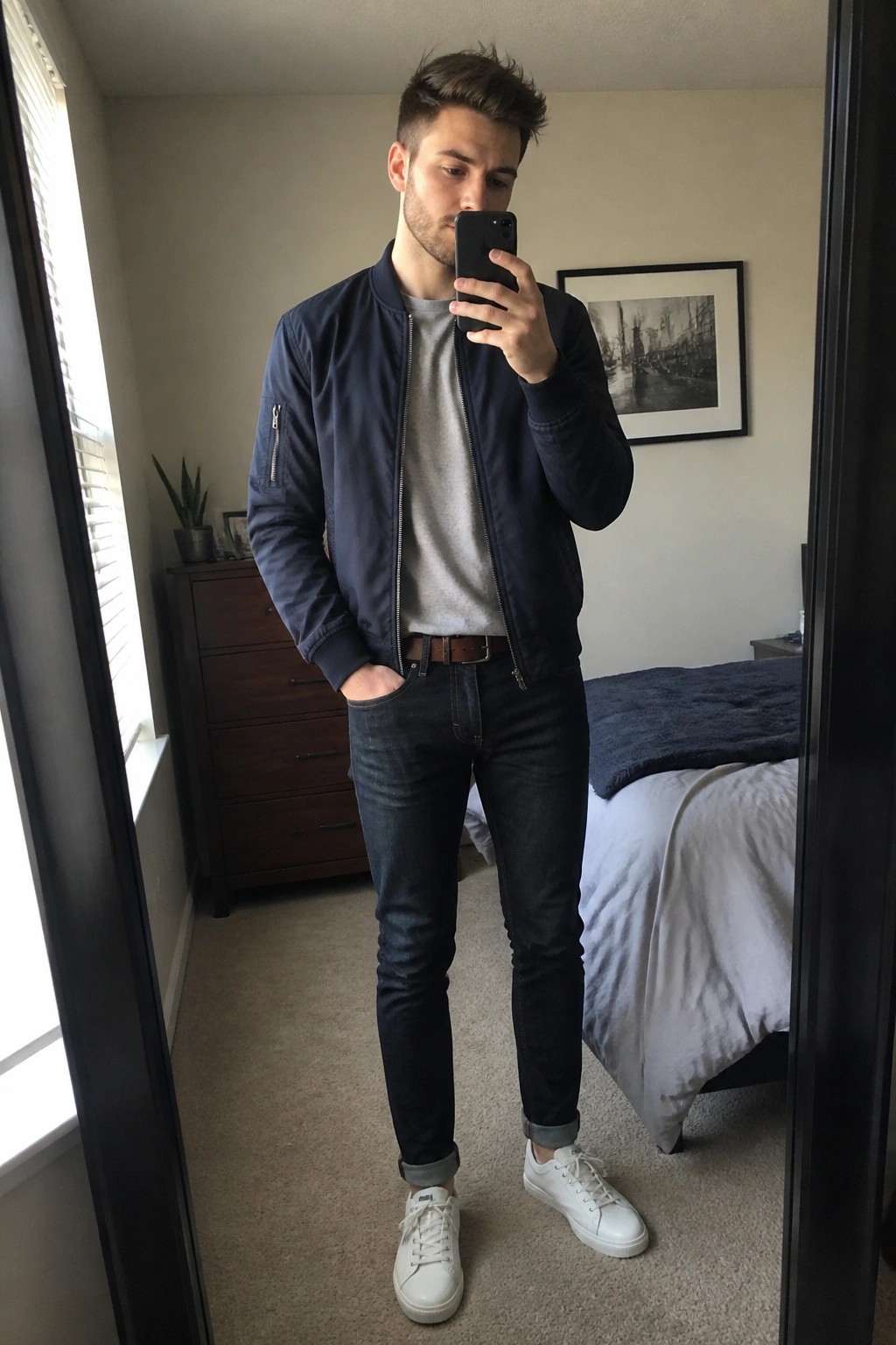 Man in navy bomber jacket taking mirror selfie in bedroom with white sneakers