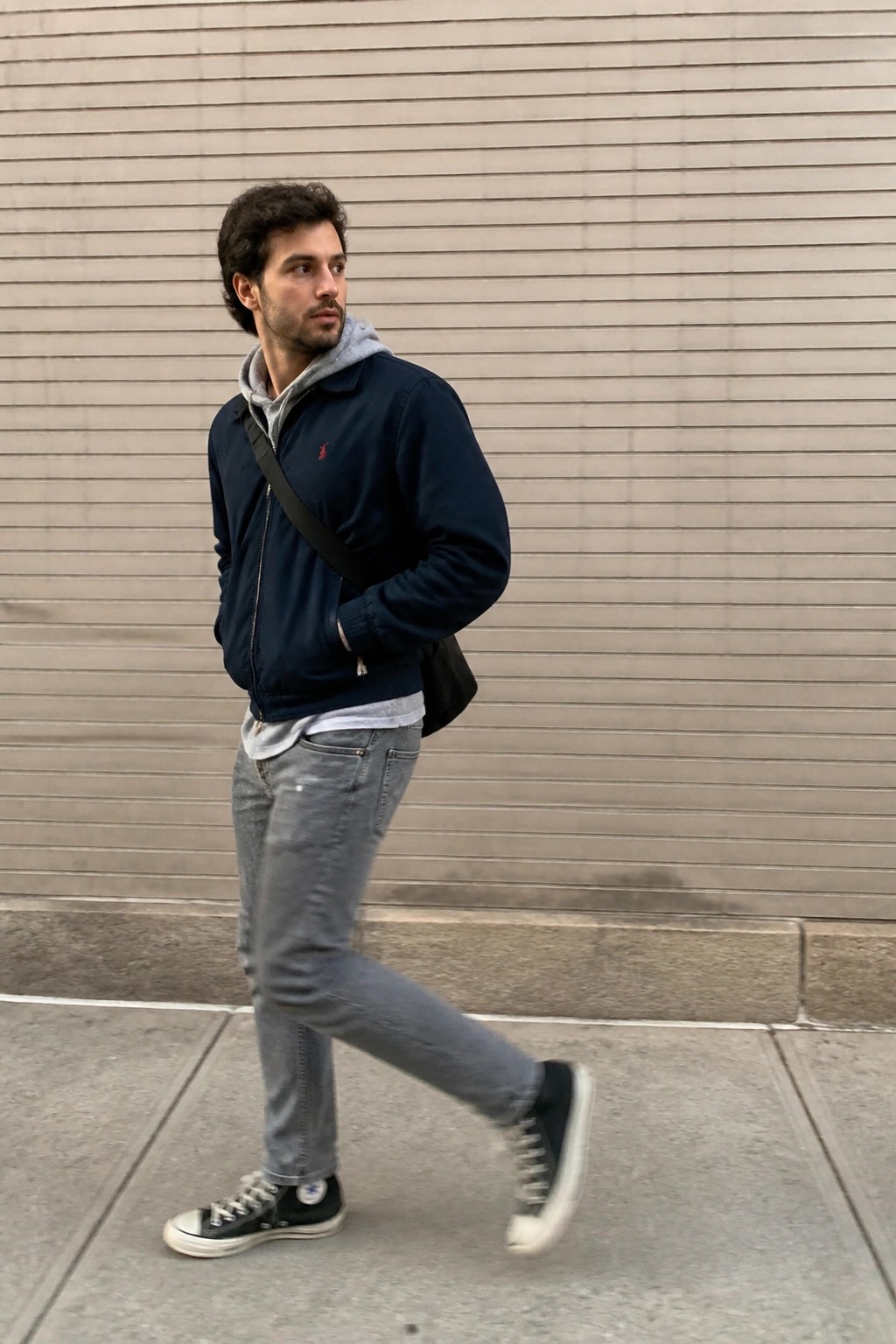 Man in navy jacket over grey hoodie and jeans walks past beige wall