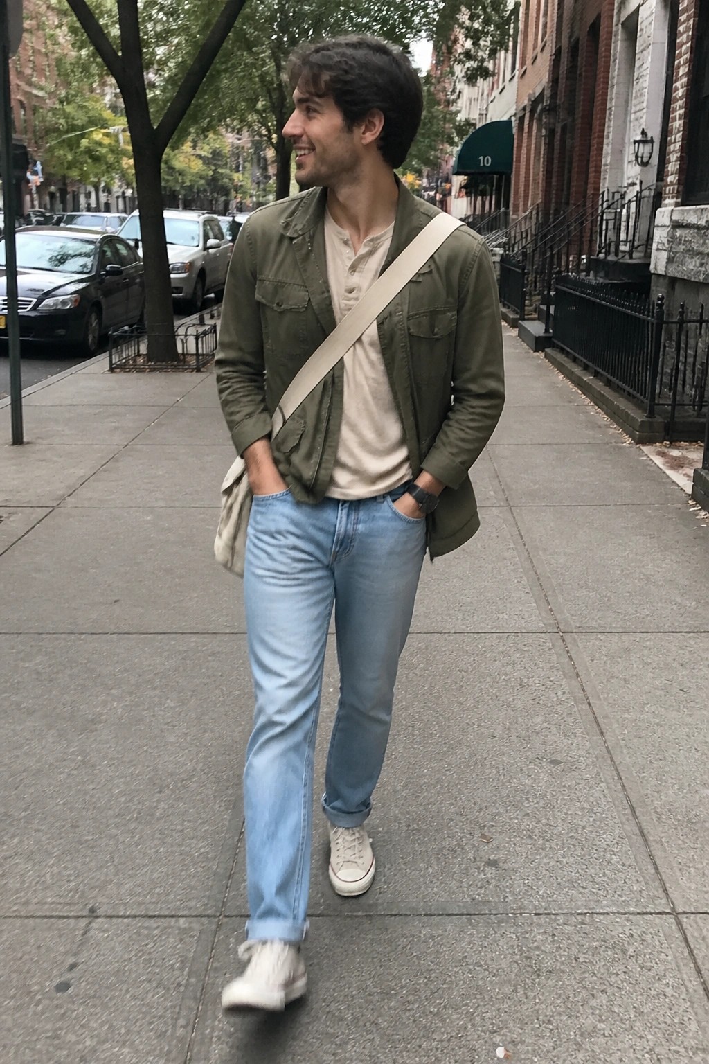Smiling man in green jacket with beige crossbody bag on city sidewalk.