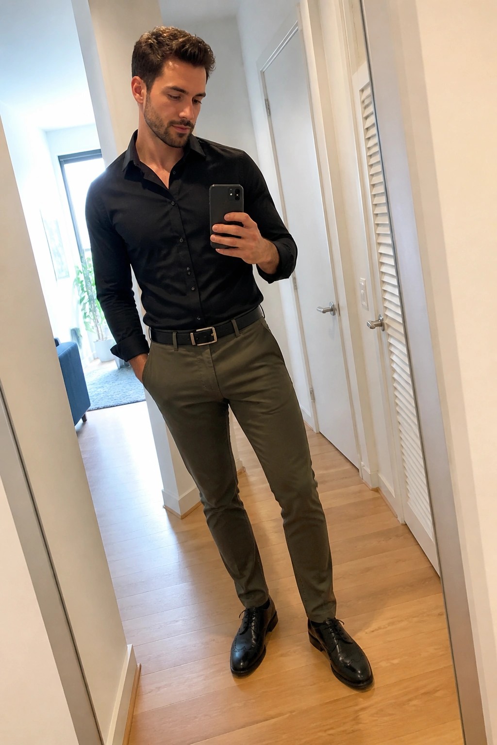 Man in black shirt and olive pants taking mirror selfie in hallway