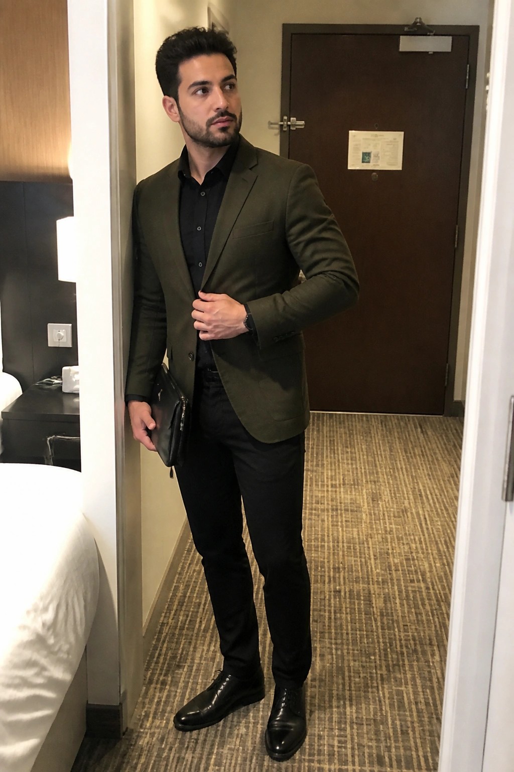 Man in olive green blazer over black shirt holding leather pouch in hotel room