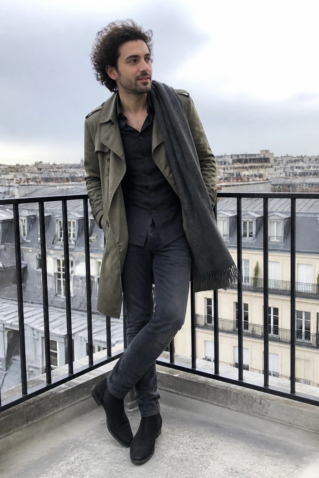 Man in olive trench coat and gray scarf on balcony overlooking city rooftops