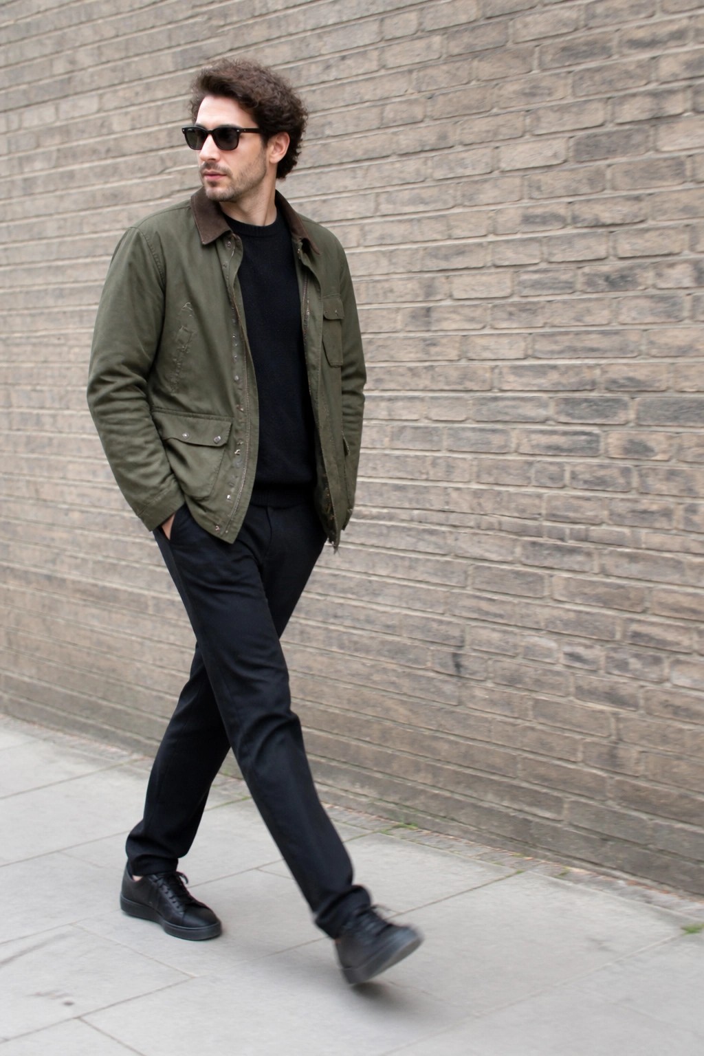 Man in green jacket over black sweater walking by brick wall