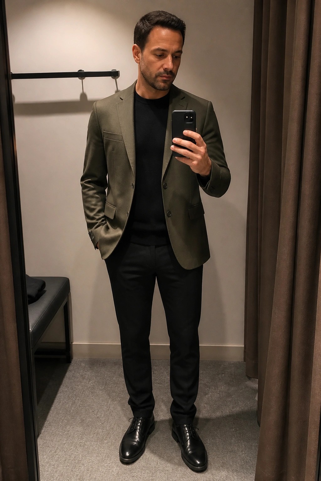 Man in olive blazer over black sweater takes mirror selfie in fitting room