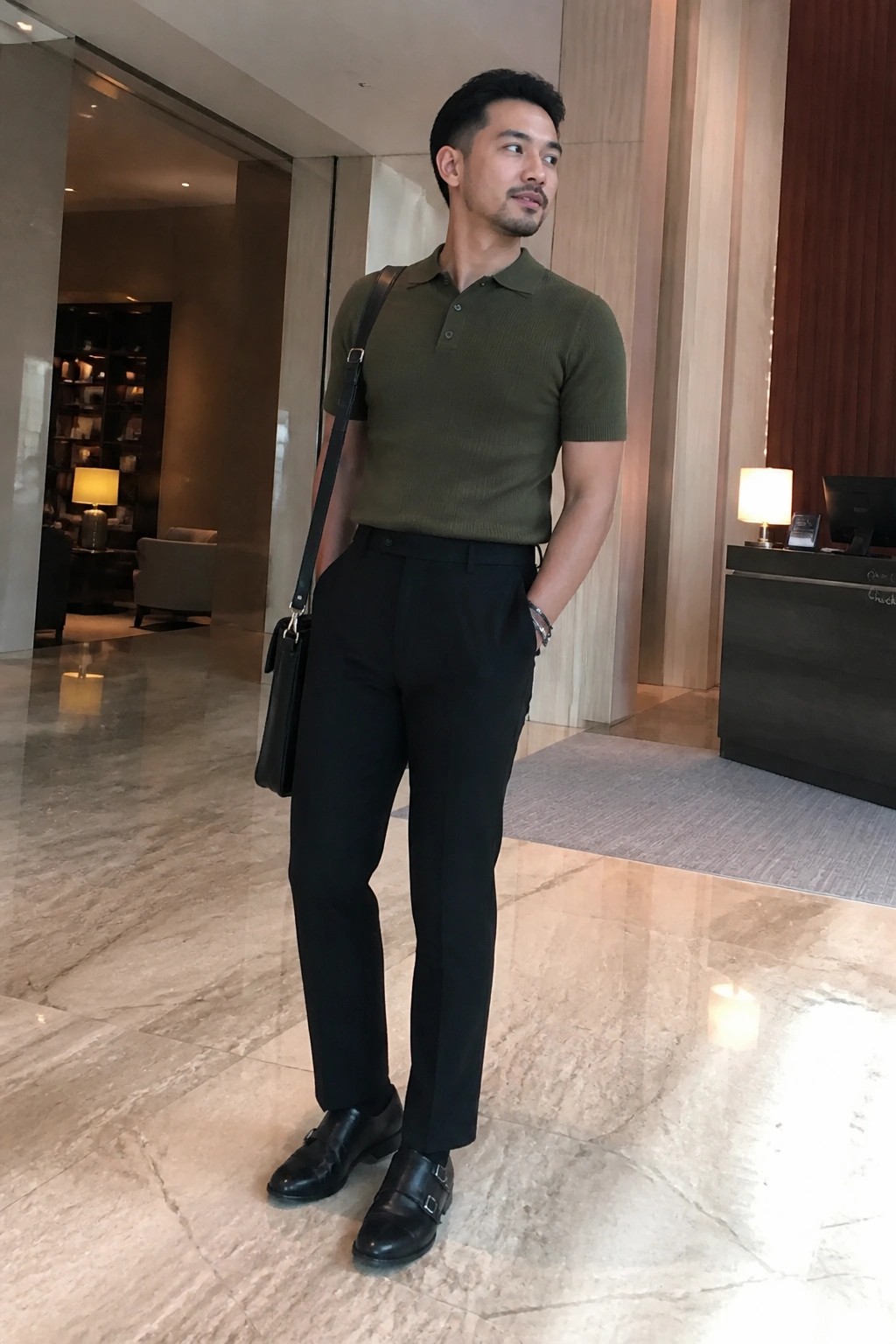 Man in olive green polo shirt and black trousers with shoulder bag in marble lobby.