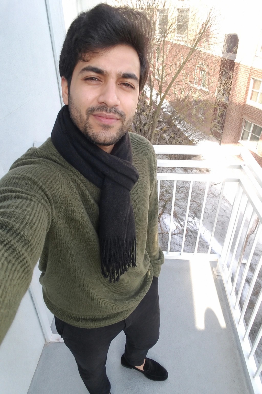 Man wearing an olive green textured knit sweater, black fringed scarf, black pants, and black slip-on shoes in a full-body selfie on a balcony.