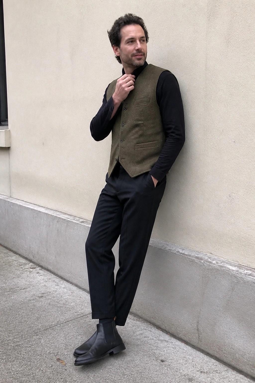 Man in olive vest over black shirt with black pants leaning on wall